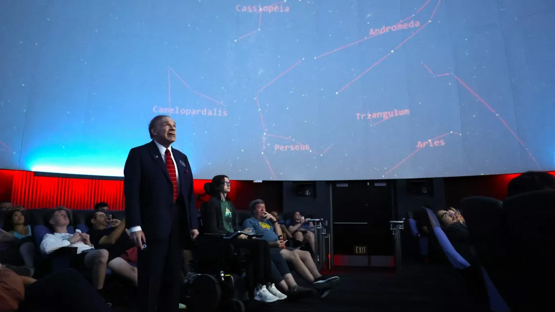 In the heart of the planetarium, the Astronomy class is about to embark on a cosmic journey. The room is dimly lit, with the vast dome above serving as a canvas for a spectacular display of stars and celestial phenomena. Students, seated in reclining chairs, are adjusting their position for optimal stargazing.