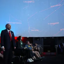 In the heart of the planetarium, the Astronomy class is about to embark on a cosmic journey. The room is dimly lit, with the vast dome above serving as a canvas for a spectacular display of stars and celestial phenomena. Students, seated in reclining chairs, are adjusting their position for optimal stargazing.